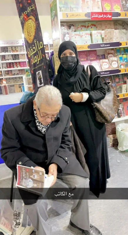 كمال رُحيّم جالسًا يكتب إهداءً في كتاب أثناء لقاء بإحدى القارئات في معرض الكتاب.