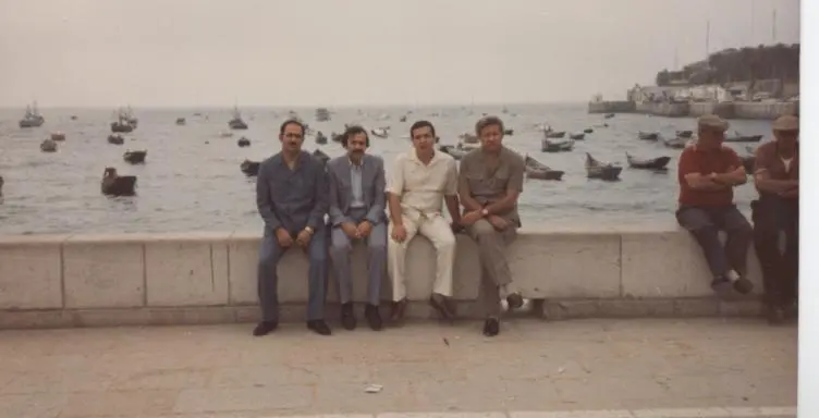 Une ancienne photo couleur de quatre hommes assis sur un muret de pierre devant une baie pleine de barques ; parmi eux Kamal Ruhayyim, en chemise et pantalon blancs.