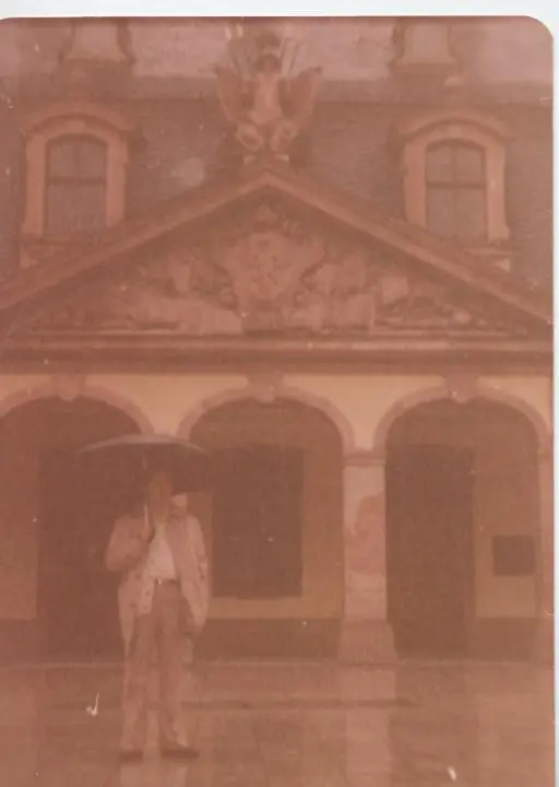 Une ancienne photo couleur de Kamal Ruhayyim tenant un parapluie un jour de pluie, devant un bâtiment baroque à arcades et fronton surmonté d’un aigle.