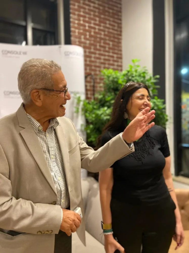 An older man in a beige jacket gestures as he talks with a smiling woman in black; plants and a brick wall behind them.