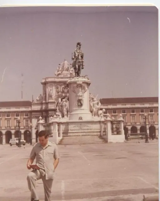 Une ancienne photo couleur de Kamal Ruhayyim en chemise et pantalon clairs, un livret à la main, devant une statue équestre sur un grand socle entourée d’arcades.