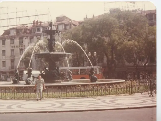 Une ancienne photo couleur de Kamal Ruhayyim devant une grande fontaine sculptée place du Rossio, avec des immeubles et un bus orange derrière.