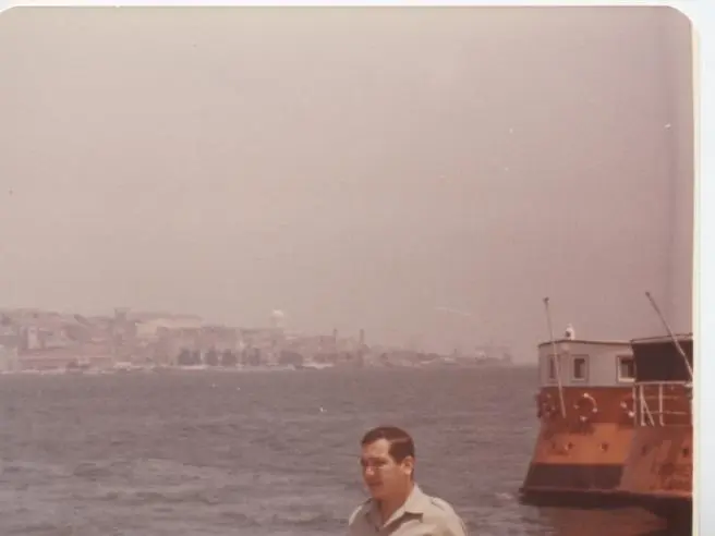Une ancienne photo couleur de Kamal Ruhayyim en chemise claire au bord du Tage, Lisbonne sur l’autre rive et un ferry orange à quai.