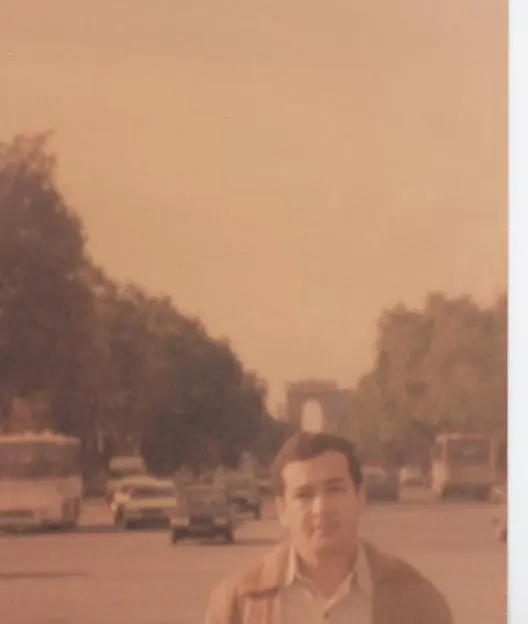 Une ancienne photo couleur : Kamal Ruhayyim au premier plan, derrière lui une large avenue arborée pleine de voitures et de bus, l’Arc de triomphe au fond.