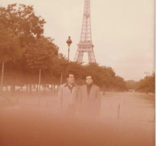 Une ancienne photo couleur de deux hommes sur une allée bordée d’arbres, la tour Eiffel derrière eux.