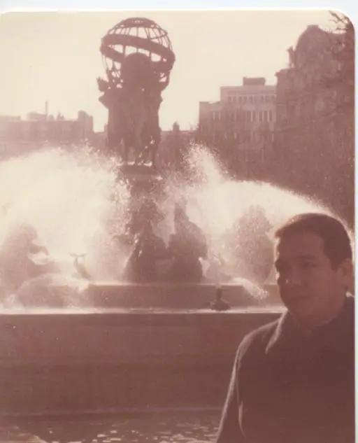 Une ancienne photo couleur : Kamal Ruhayyim au premier plan, derrière lui une fontaine jaillissante surmontée de statues portant un globe.