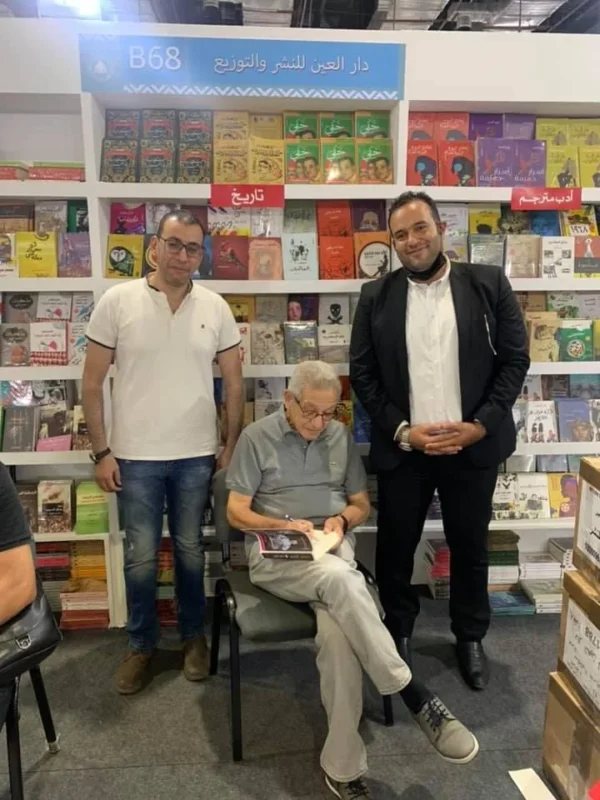 رجل مسنّ جالس يوقّع كتابًا في جناح دار نشر، ويقف بجواره رجلان، وخلفهم رفوف كتب ملونة.