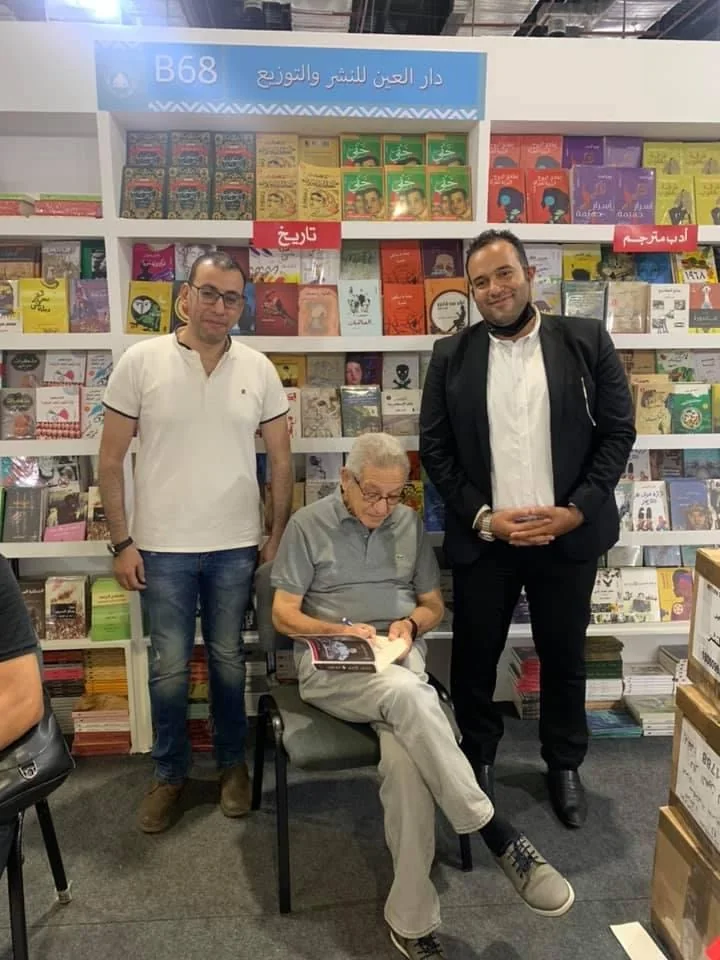 رجل مسنّ جالس يوقّع كتابًا في جناح دار نشر، ويقف بجواره رجلان، وخلفهم رفوف كتب ملونة.