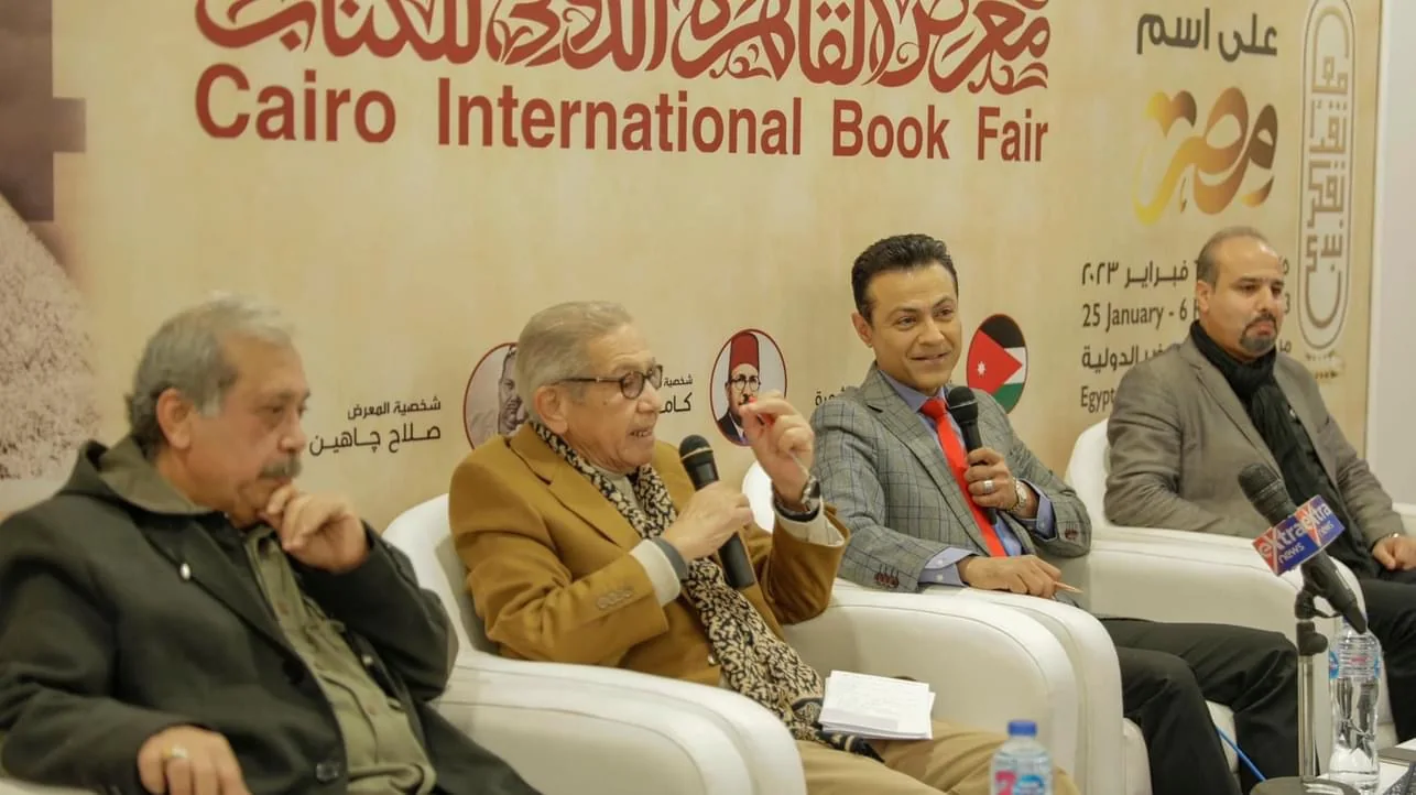 Kamal Ruhayyim speaking into a microphone at a Cairo International Book Fair panel with fellow participants.