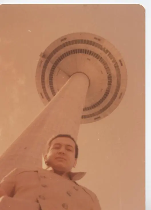 Une ancienne photo couleur prise en contre-plongée : Kamal Ruhayyim en manteau, sous une haute tour de béton surmontée d’une plateforme ronde.