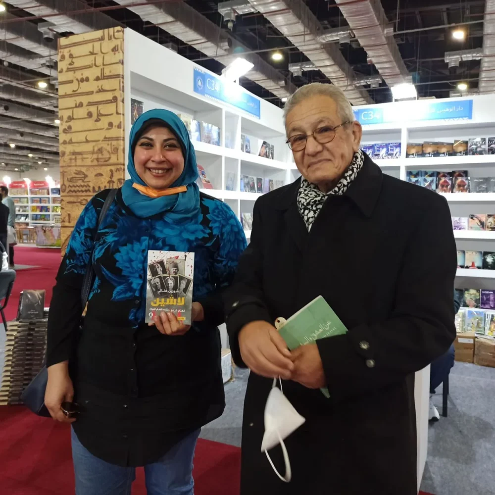 Eine Frau mit blauem Kopftuch und einem Buch steht neben einem älteren Mann in dunklem Mantel und Schal, der ein kleines grünes Buch hält, in einem Gang der Buchmesse.