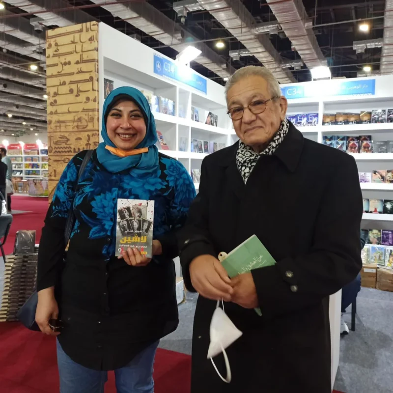امرأة بحجاب أزرق تحمل كتابًا تقف بجوار رجل مسنّ بمعطف داكن ووشاح يحمل كتابًا أخضر صغيرًا، في أحد ممرات معرض الكتاب.