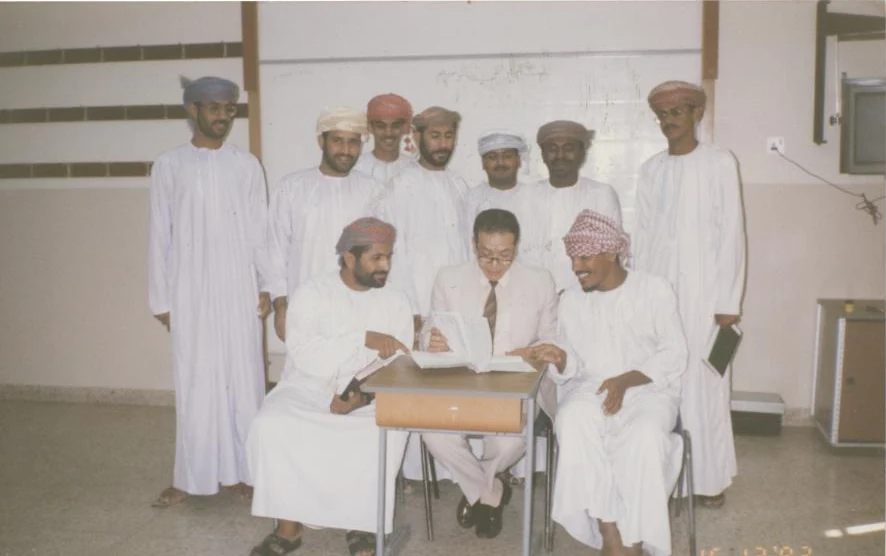 Kamal Ruhayyim sitzt mit einem aufgeschlagenen Buch an einem Tisch, umgeben von seinen Studenten in omanischer Kleidung an der Polizeiakademie in Oman.