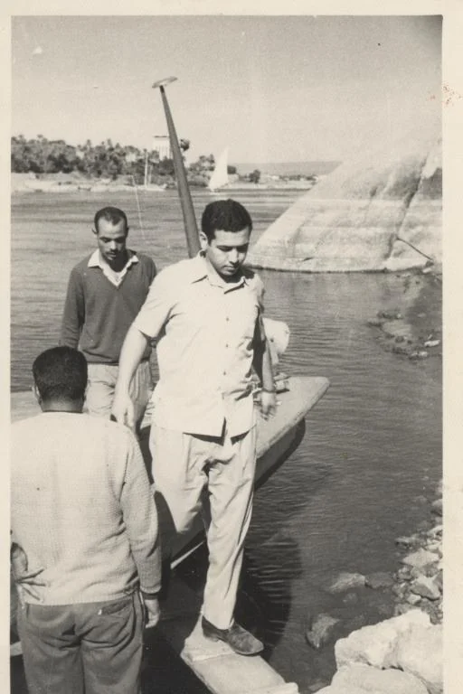 Photographie en noir et blanc d’hommes près d’une petite embarcation au bord du fleuve.